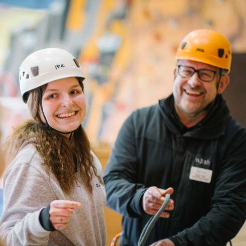 Young woman smiling in front of a climbing wall with an instructor behind her