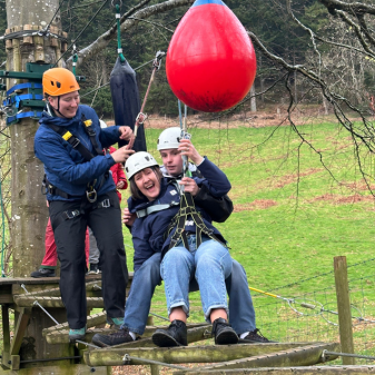 Woman wobbling on high ropes course supported by a man behind her and an instructor reaching to help