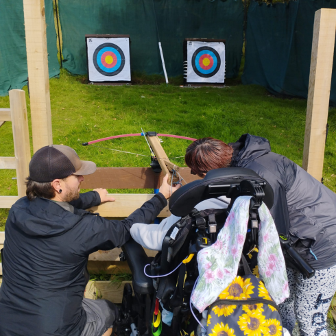 Instructor helping a wheelchair user use a crossbow to fire arrows are targets in the distance