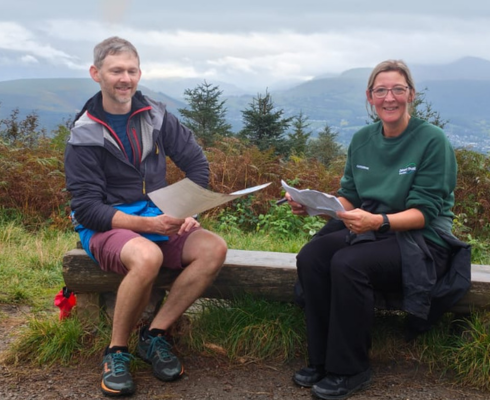 Two people reading maps on a bench with a view of mountains and clouds behind