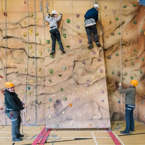 Two climbers high on an indoor climbing wall with two instructors stood below