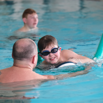 Young boy in goggles using a pool noodle supported by his dad in the swimming pool
