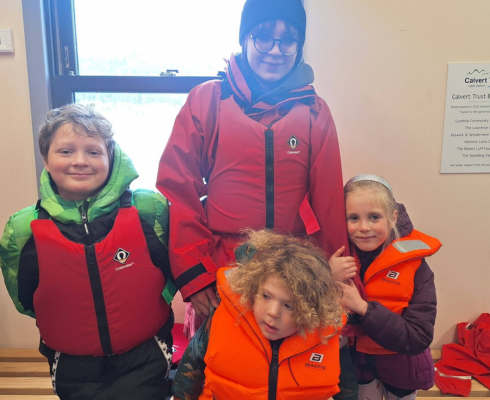 Woman and three children in waterproofs and buoyancy aids in the boathouse before canoeing