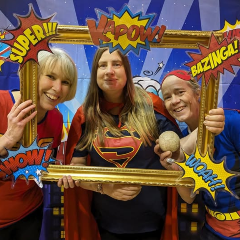 Three women in superhero attire holding a photo frame