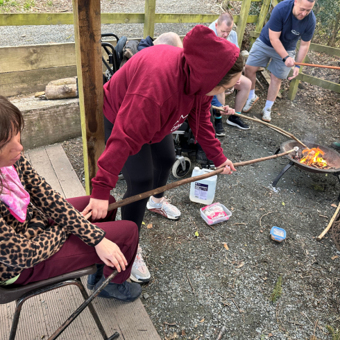 Woman supporting another holding a long stick over a fire pit with a marshmallow on