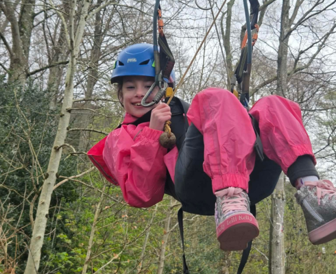 Girl in pink waterproofs hanging in a zip wire harness smiling having been down the zip wire