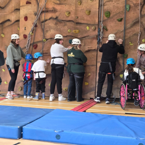 Group of mixed ability adults and children against a climbing wall with harnesses on