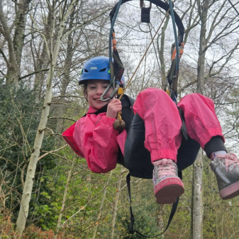 Girl in pink waterproofs hanging in a zip wire harness smiling having been down the zip wire
