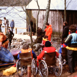 Group of people, some in wheelchairs, outside a boathouse and jetty with the lake in the background preparing to go onto the water in canoes
