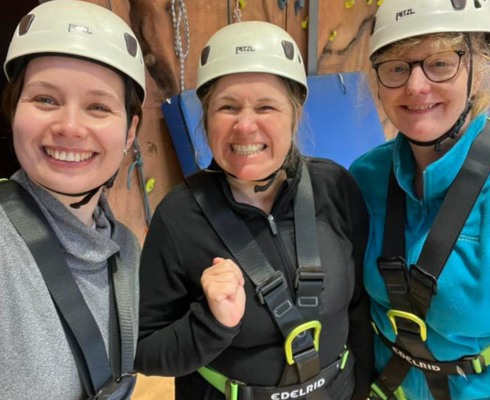 Three women smiling in front of an indoor climbing wall wearing harnesses and helmets
