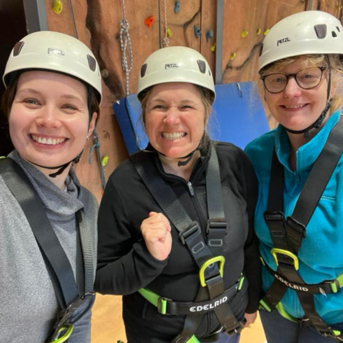 Three women smiling in front of an indoor climbing wall wearing harnesses and helmets