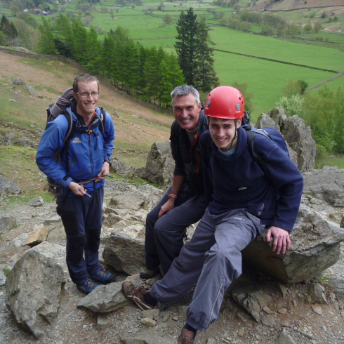 Three people sat on rocks at top of cliff face with valley behind and below