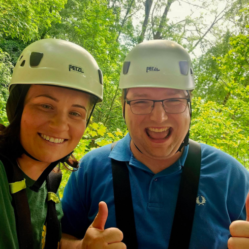 Man and woman wearing helmets smiling with thumbs up with trees in the background