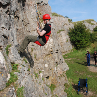 Man climbing up crag face with wheelchair below