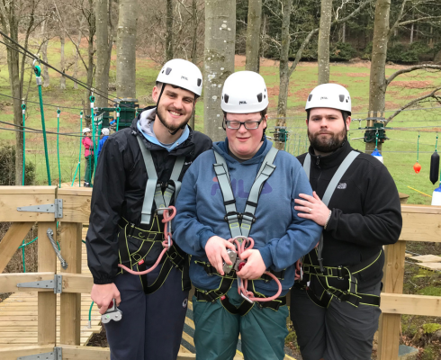 Three men smiling in helmets and harness in front of a high ropes course